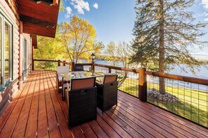 Terraza de madera con una mesa y sillas, con vista a un lago rodeado de árboles y un cielo despejado.