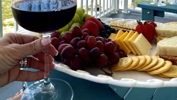 Een balkon met een eettafel, compleet met een glas rode wijn, een bord vol fruit, kaas, crackers en sandwiches.