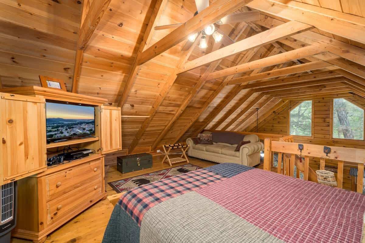 A cozy attic bedroom with a wooden ceiling, a television, a bed, and a window overlooking a forest.