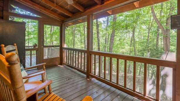 A covered porch with wooden flooring, a ceiling fan, and a view of a forest.