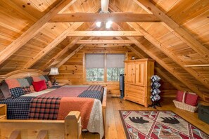 Un dormitorio acogedor en un ático con techo de madera, una cama con ropa de cama escocesa, una ventana y un aparador con revistas.