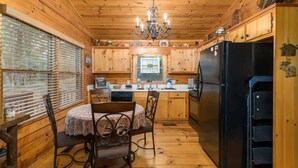 Interior de una cabaña de madera con una mesa de comedor, sillas, una araña y un refrigerador negro.