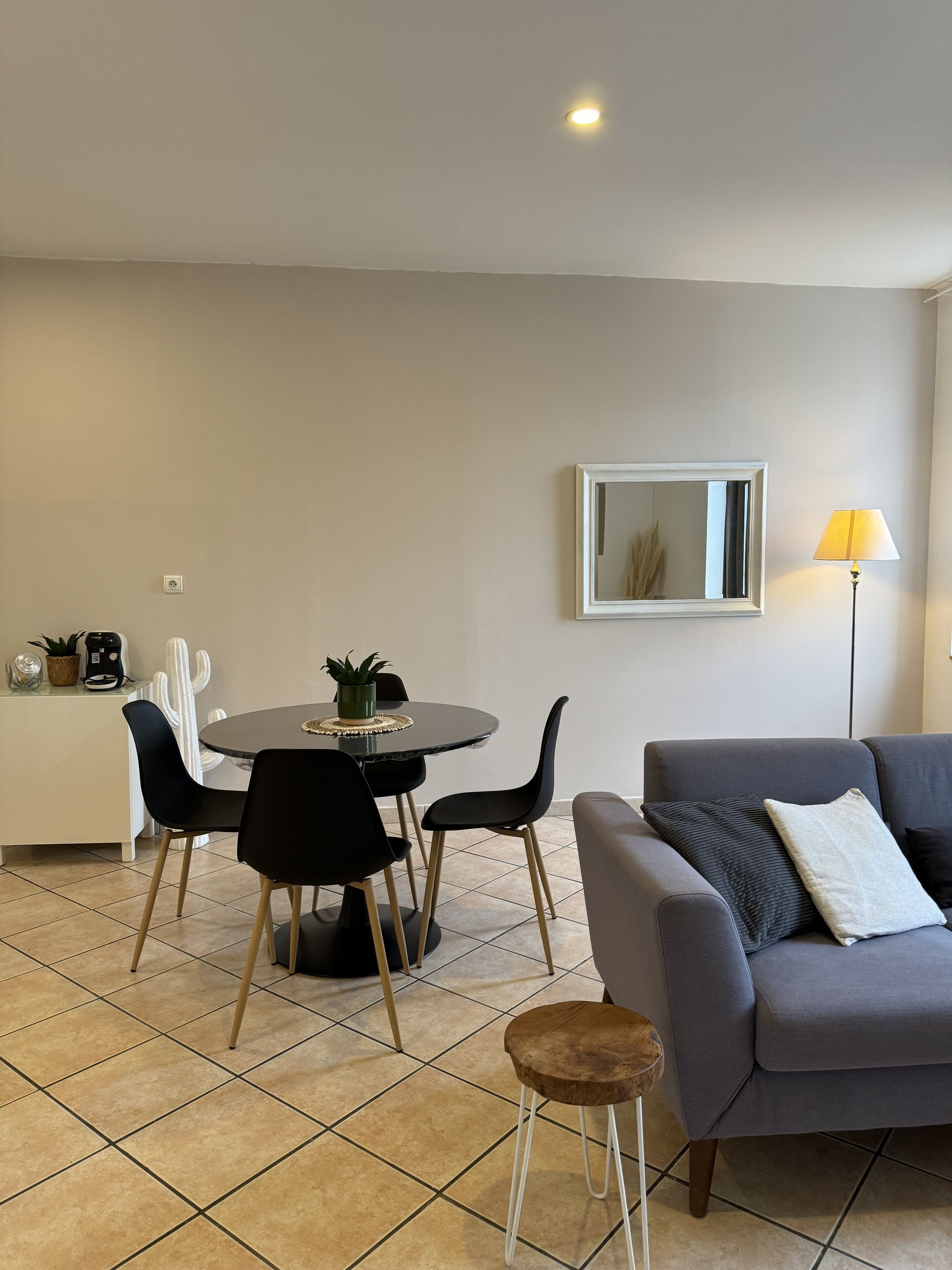Ein modernes Wohnzimmer mit einem grauen Sofa, einem runden Esstisch mit schwarzen Stühlen, einem kleinen Holzschemel, einer Kakteenpflanze und einer Lampe.