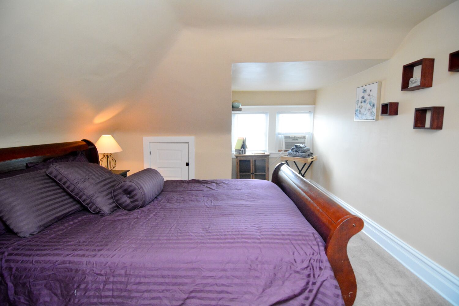 A bedroom with a large bed, a nightstand with a lamp, a door, and a small table with a chair.