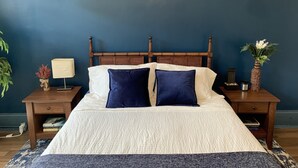 Una camera da letto con testiera in legno, due comodini, un letto con biancheria bianca e blu navy, una lampada, un vaso con fiori e una pianta.