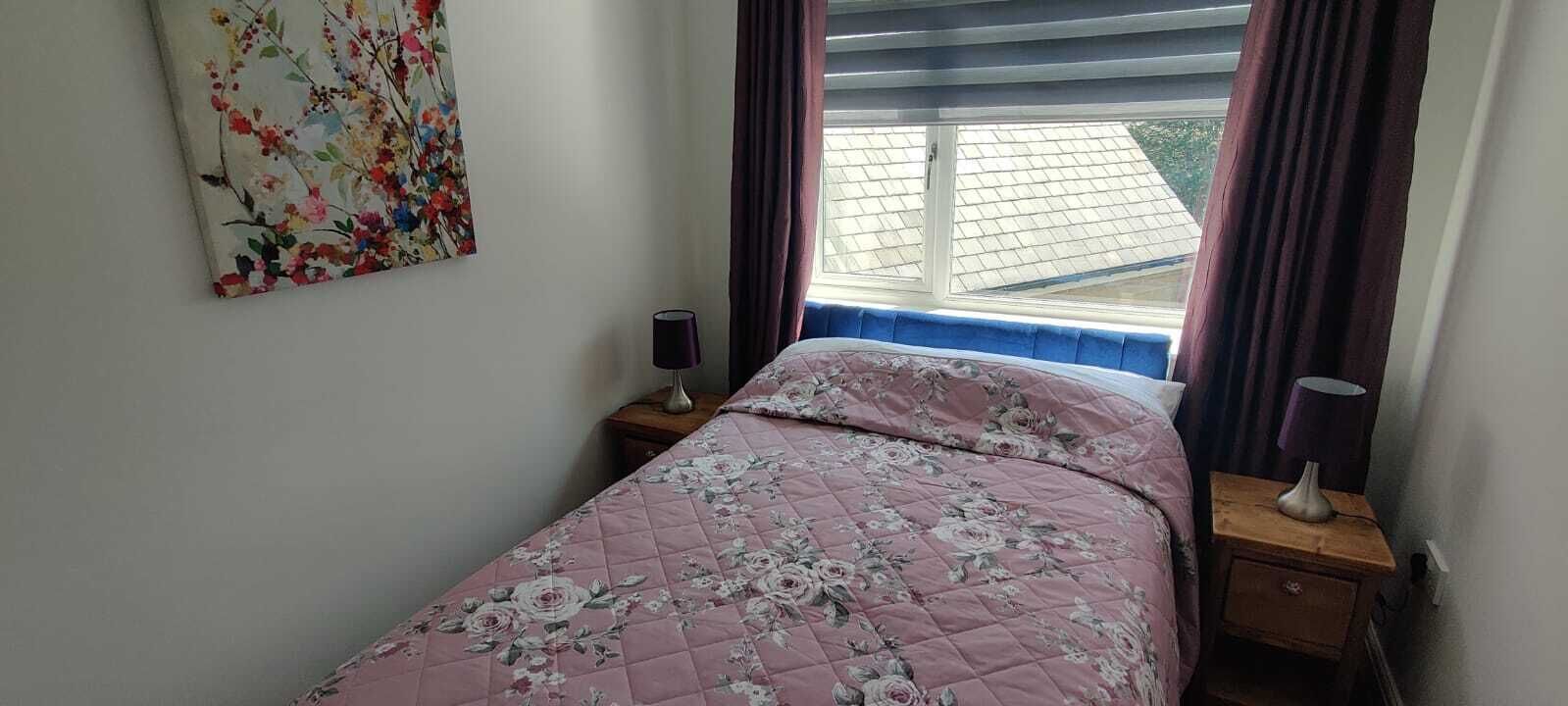 Ein Schlafzimmer mit einem Bett, zwei Nachttischen, einem Fenster mit Vorhängen und einem Blumenbild an der Wand.
