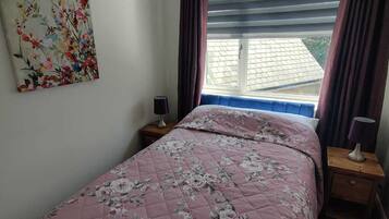 Ein Schlafzimmer mit einem Bett, zwei Nachttischen, einem Fenster mit Vorhängen und einem Blumenbild an der Wand.