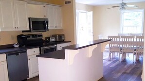 A kitchen with white cabinets, a microwave, a stove, and a dishwasher. There is a dining area with a table and chairs. - Bar Harbor Serenity close to Hadley Point Beach (Bar Harbor)