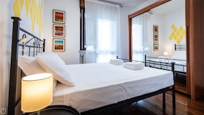 Een keurig opgemaakt bed met witte lakens, een bedlampje en een raam met dunne gordijnen.