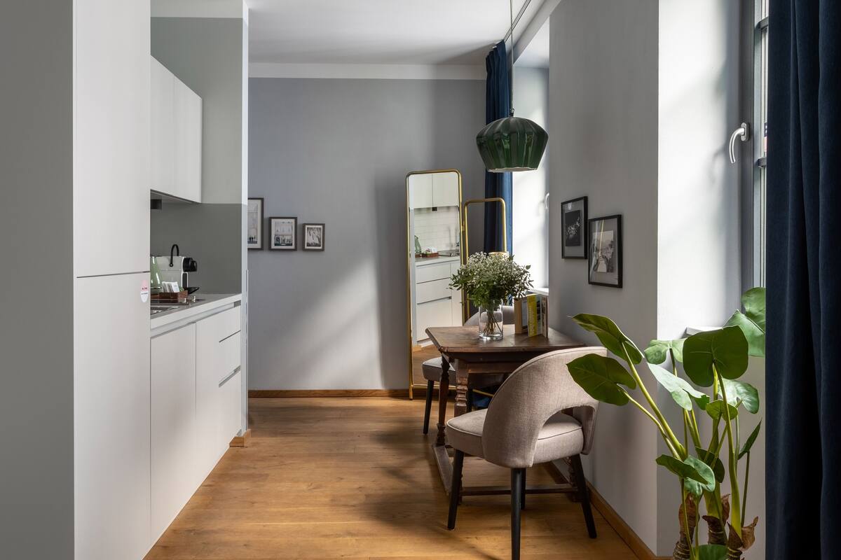 A modern kitchen with white cabinets, a wooden floor, a dining table with chairs, a mirror, and a potted plant.