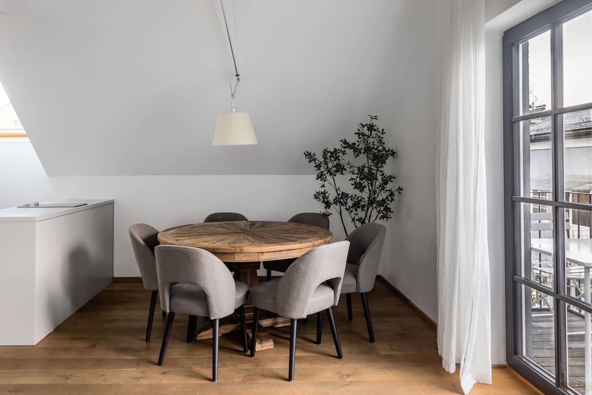 A dining area with a round wooden table, six grey chairs, a hanging light fixture, a potted plant, and a large window with curtains.