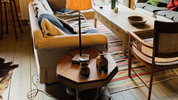 A cozy cabin interior with a wooden ceiling, a sofa, a table, and a view of mountains through a large window.