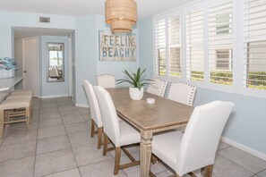 A dining area with a wooden table, white upholstered chairs, a potted plant, and a 'Feelin' Beachy' sign on the wall.