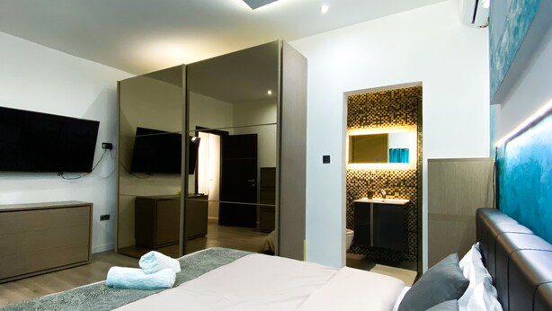Habitación de hotel moderna con una cama grande, televisión, espejo y baño visible a través de una puerta abierta.