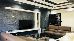 Una sala moderna con un televisor de pantalla plana montado en la pared, un sofá de cuero marrón, un sillón blanco y una mesa de centro de vidrio.