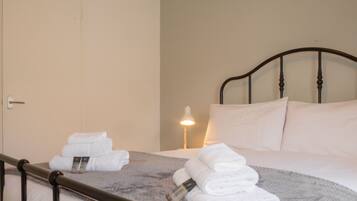 A neatly made bed with white linens and a gray blanket, a bedside lamp, and a door in the background.