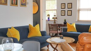 A living room with a blue sofa, yellow cushions, a round coffee table, and a dining area with a table set for two. - Sydney Wharf House (Bath)