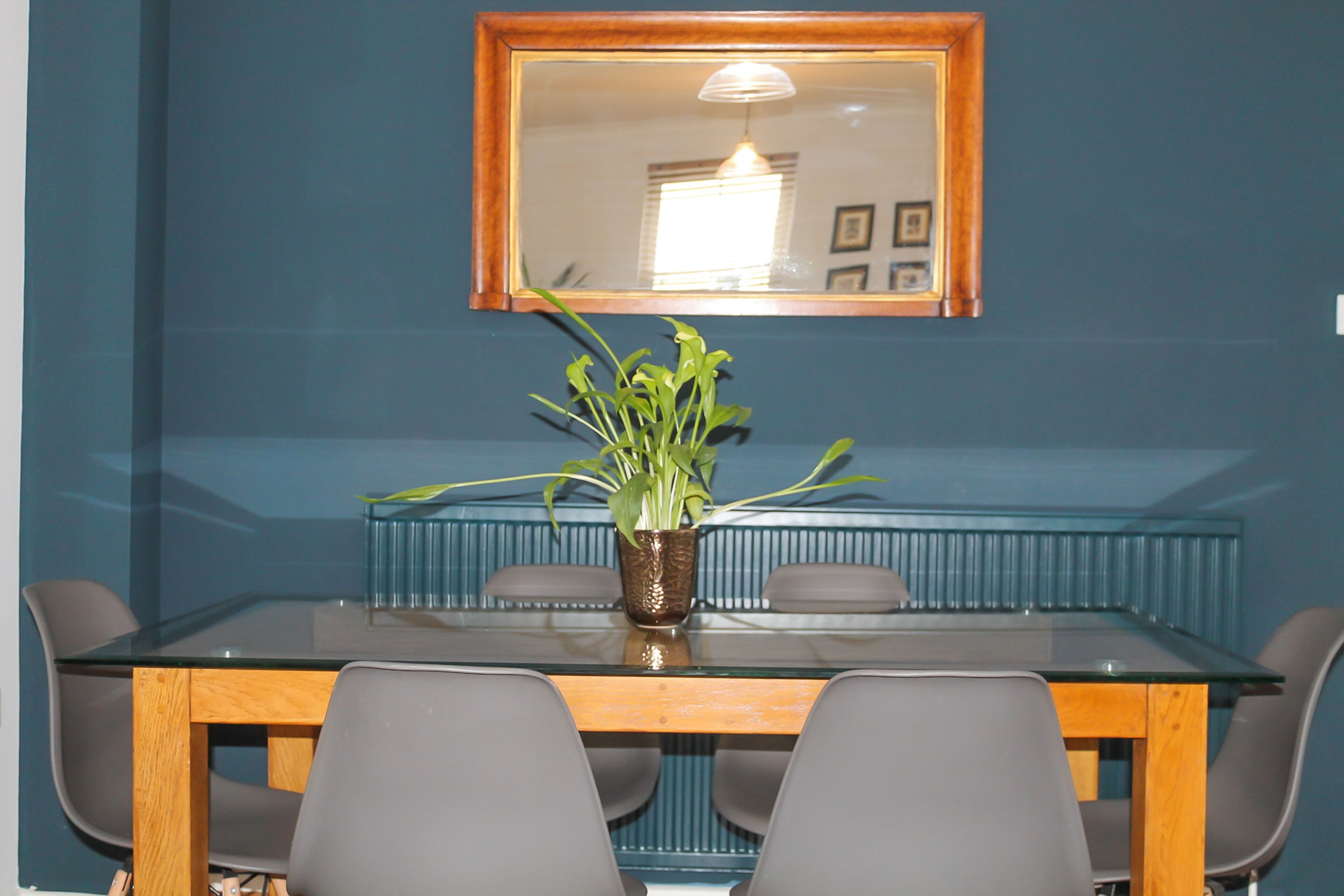Een eetgedeelte met een tafel met glazen blad, vier grijze stoelen, een houten bank en een pot met plant.