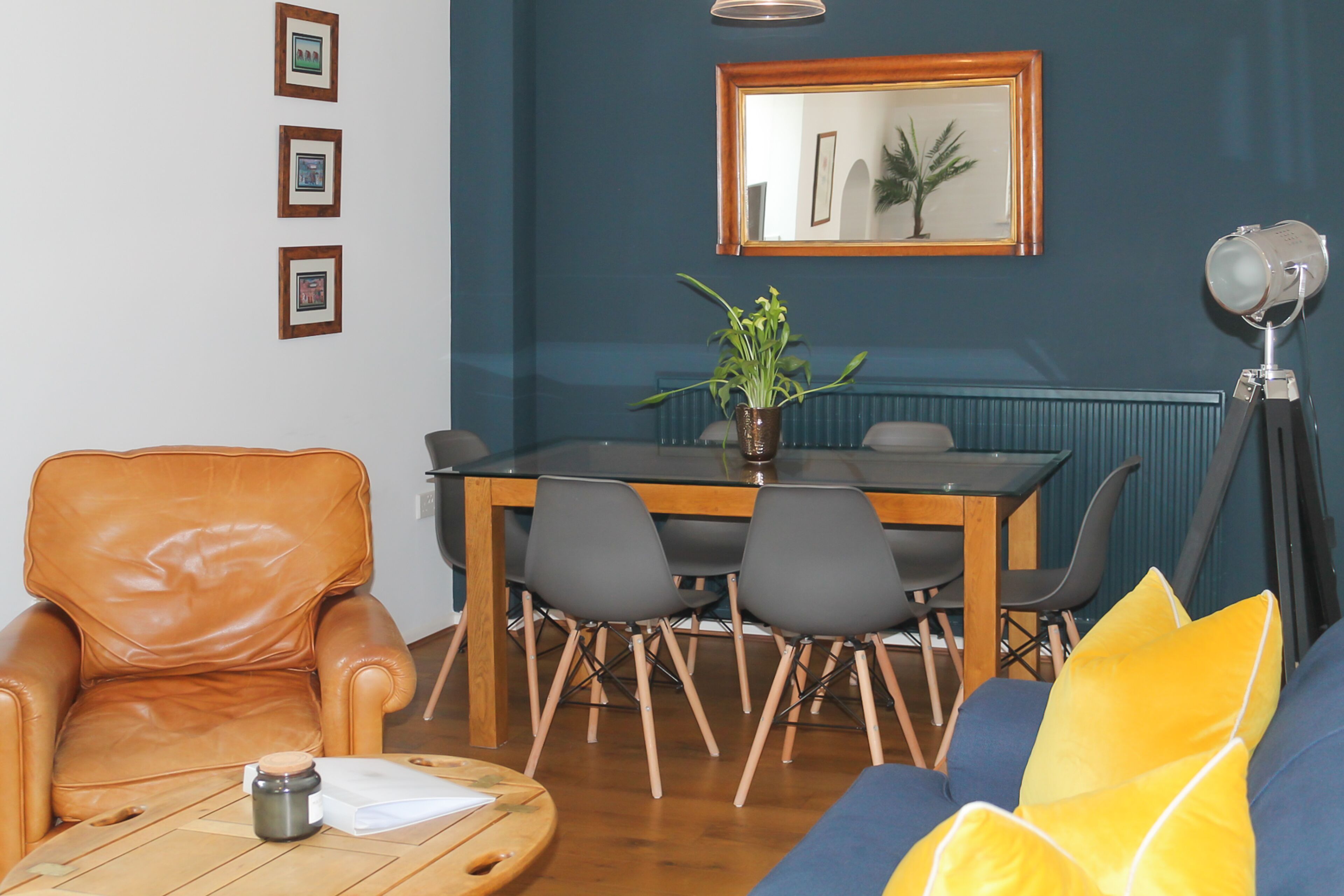 Una sala de estar con un sillón de cuero marrón, una mesa de centro de madera, un sofá azul con cojines amarillos, una mesa de comedor con sillas y una planta en maceta.