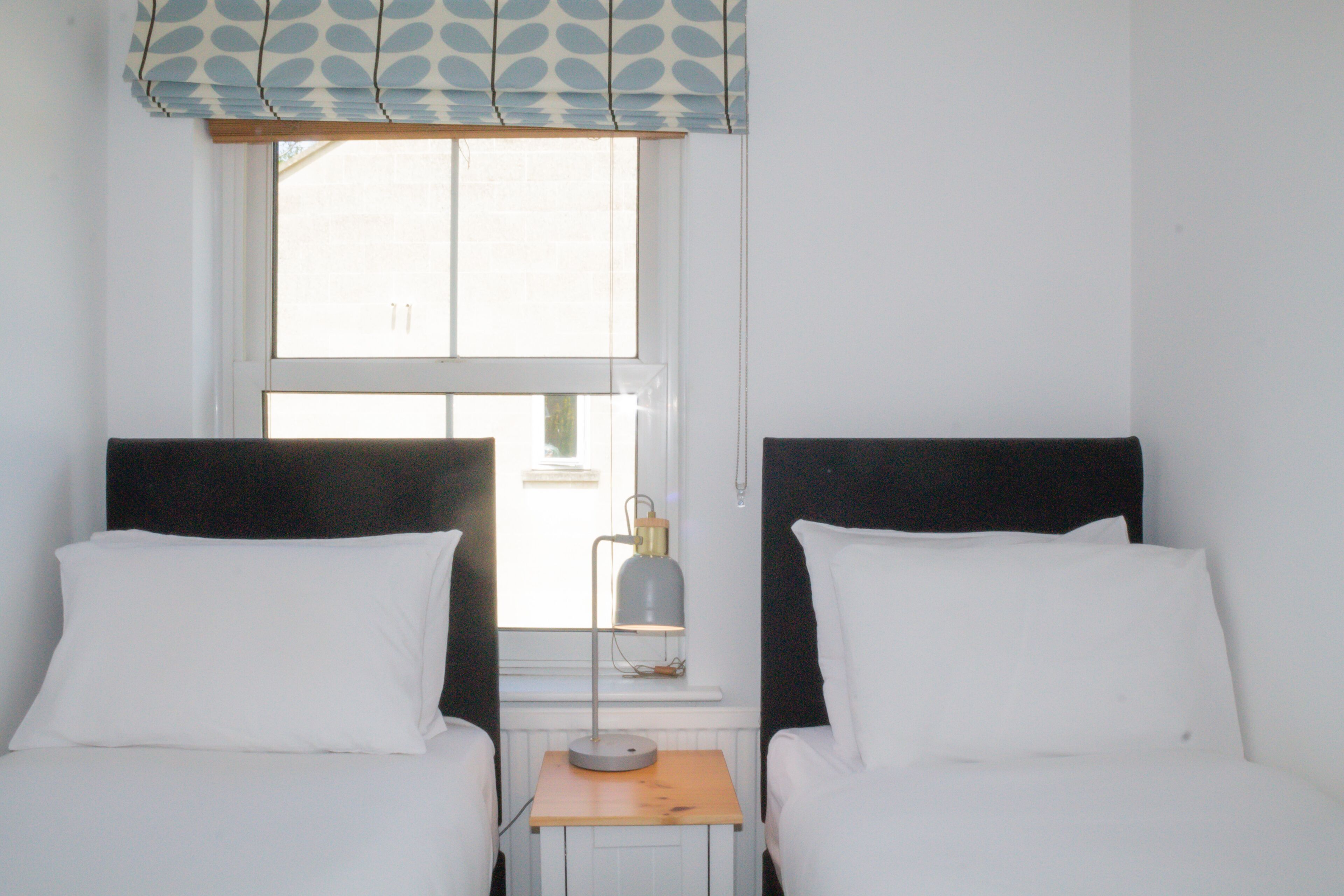 A room with two beds, a small table with a lamp, and a window with blinds.
