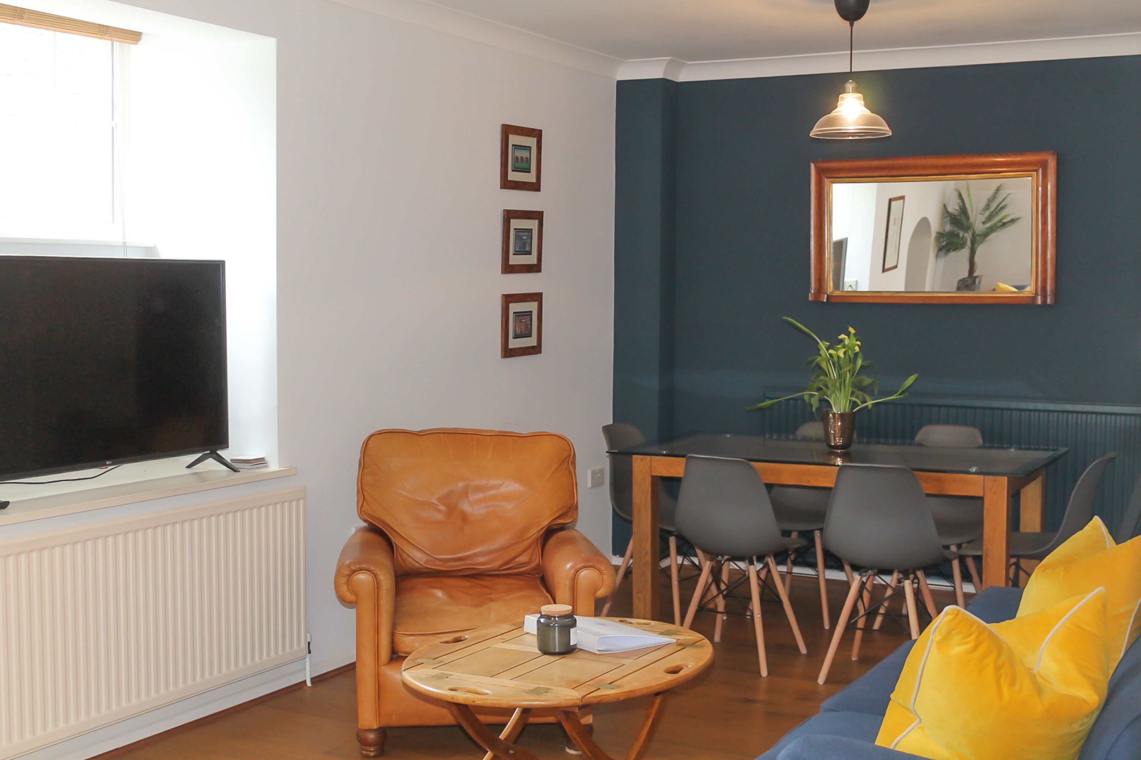 A living room with a leather armchair, a round coffee table, a blue sofa with yellow cushions, a dining table with chairs, a TV on a stand, and a mirror on the wall.