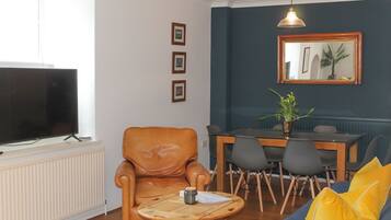 A living room with a leather armchair, a round coffee table, a blue sofa with yellow cushions, a dining table with chairs, a TV on a stand, and a mirror on the wall.