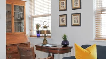 Een kamer met een houten bureau, een stoel, een blauwe bank met een geel kussen en botanische kunstwerken in een lijst aan de muur.