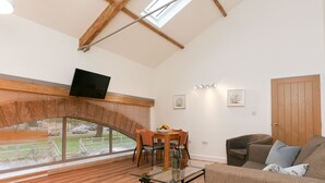Un vaste espace de vie doté d’un haut plafond, de poutres apparentes, d’une table à manger avec des chaises, d’un canapé et d’un téléviseur fixé au mur.