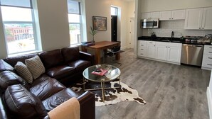 Un salon moderne avec un canapé en cuir, une table basse en verre et un coin repas comprenant une table et des chaises en bois. On aperçoit à l’arrière-plan une cuisine avec des meubles blancs et des appareils électroménagers en acier inoxydable.