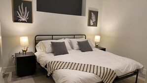 Un lit bien fait, avec du linge de lit blanc et un plaid à rayures, deux lampes de chevet et des œuvres d'art représentant des fougères, encadrées, accrochées aux murs.