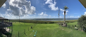 A grassy area with a wooden deck, a palm tree, and a view of a body of water. - First row Gulf views! (Bolivar Peninsula)