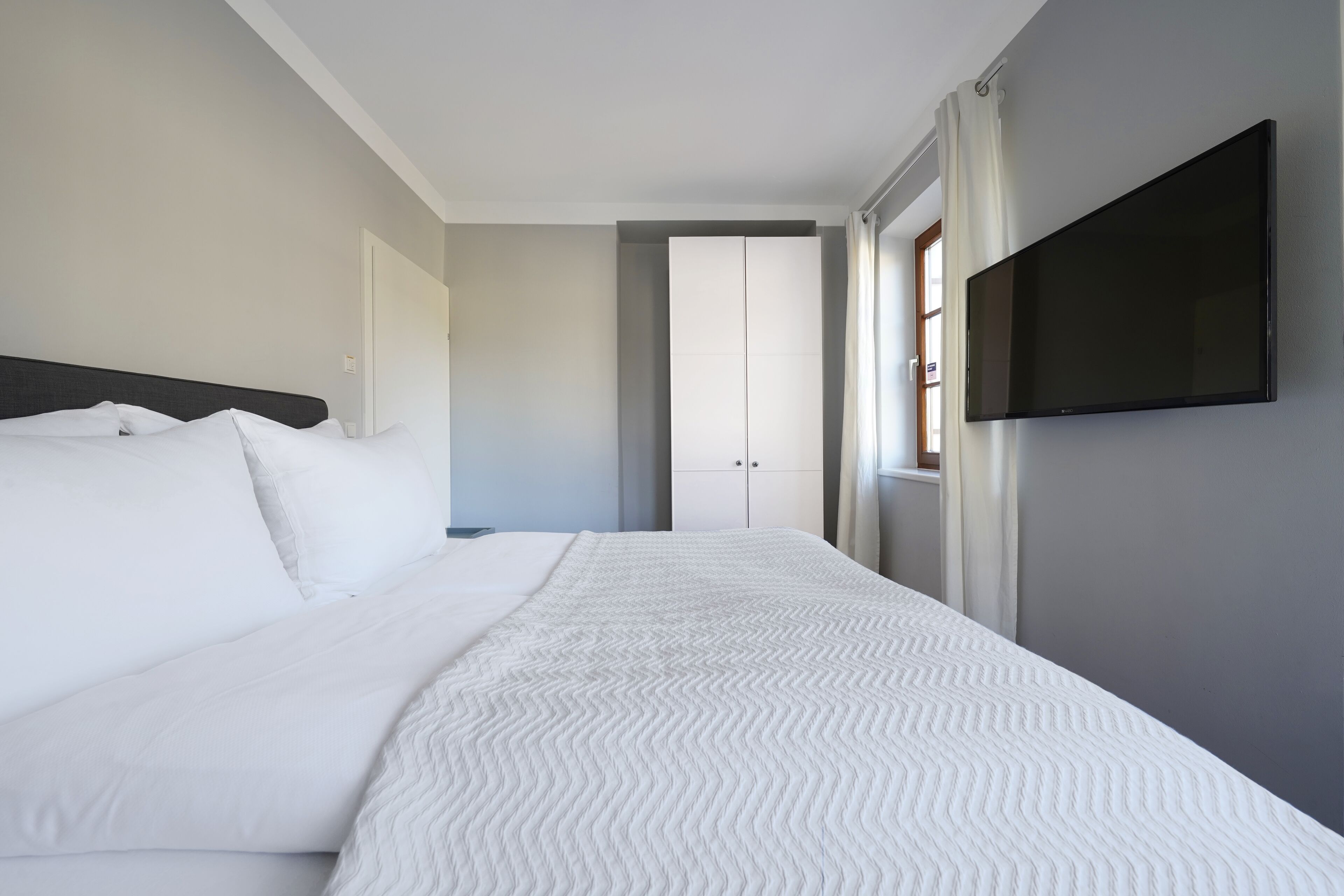 A modern hotel room with a neatly made bed, a flat-screen TV mounted on the wall, and a door leading to another room.