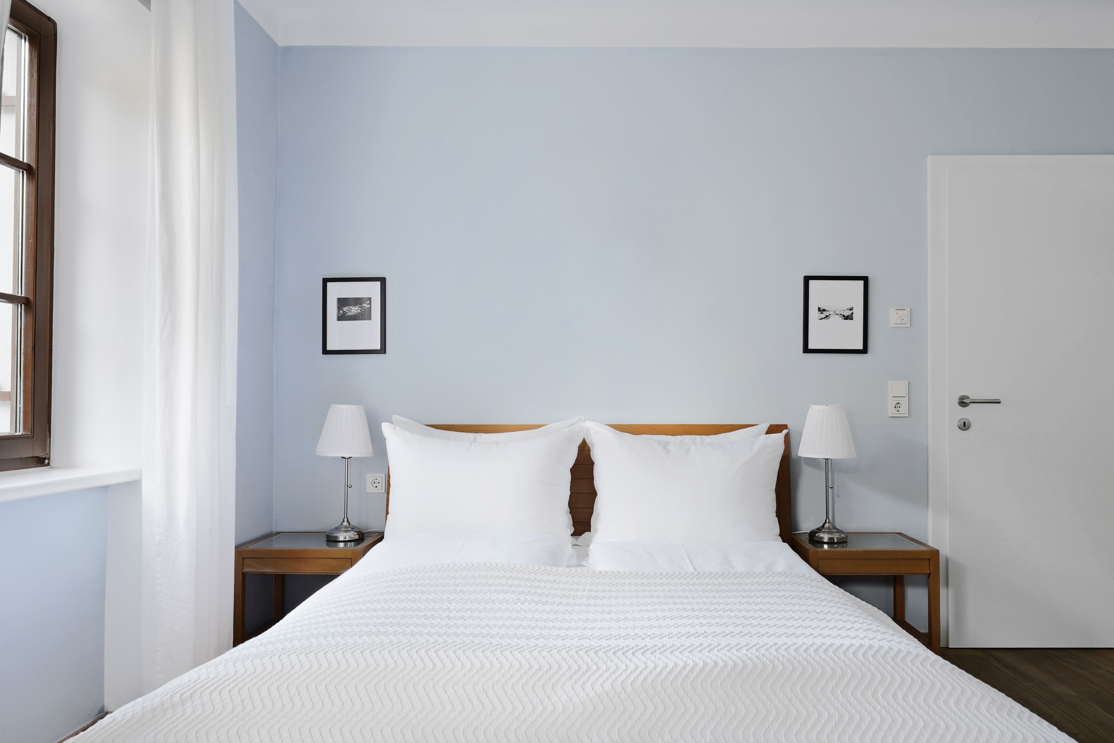 A neatly made bed with white linens, two bedside tables with lamps, and two framed pictures on the wall.