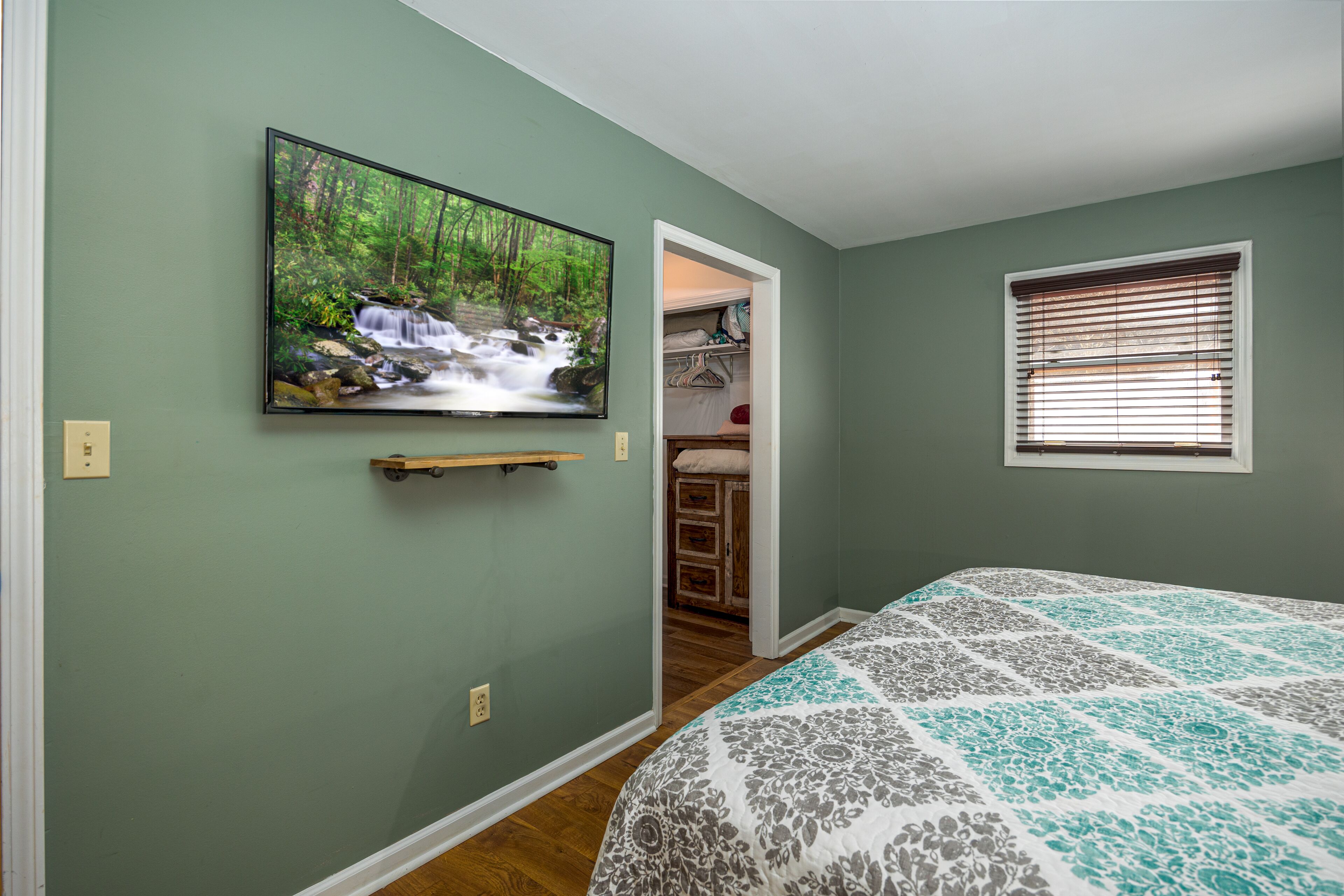 A bedroom with a bed, a TV mounted on the wall, a closet, and a window with blinds.