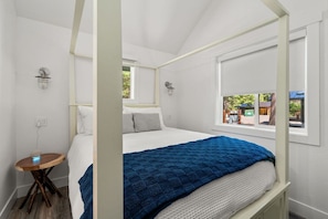 A bedroom with a four-poster bed, a bedside table with a lamp, and a window with a view of the outside.