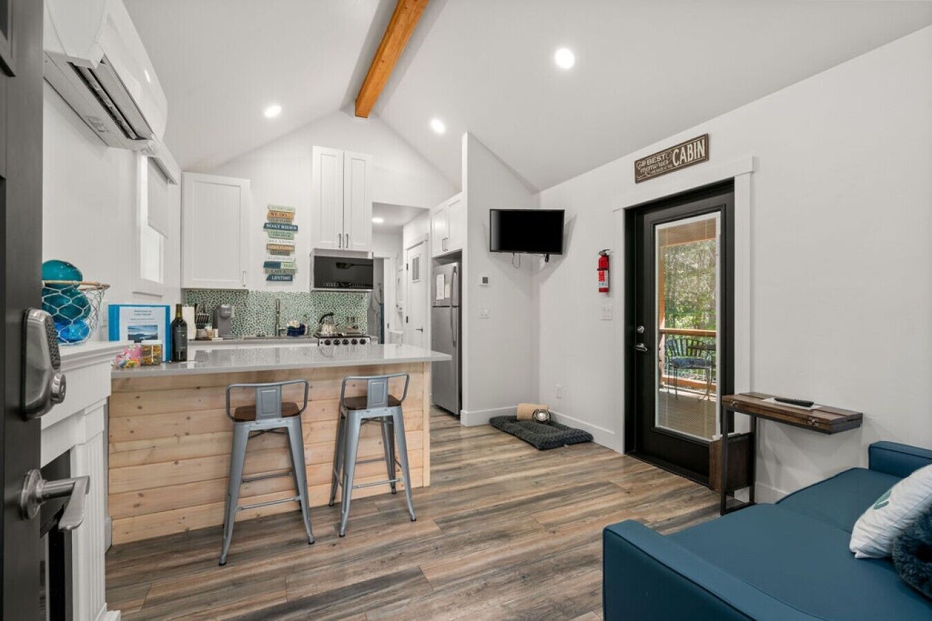 A compact kitchen with a central island, bar stools, a TV mounted on the wall, and a sliding door leading to an outdoor area.
