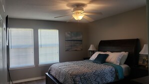 A bedroom with a bed, ceiling fan, two windows with blinds, and a nightstand with a lamp.