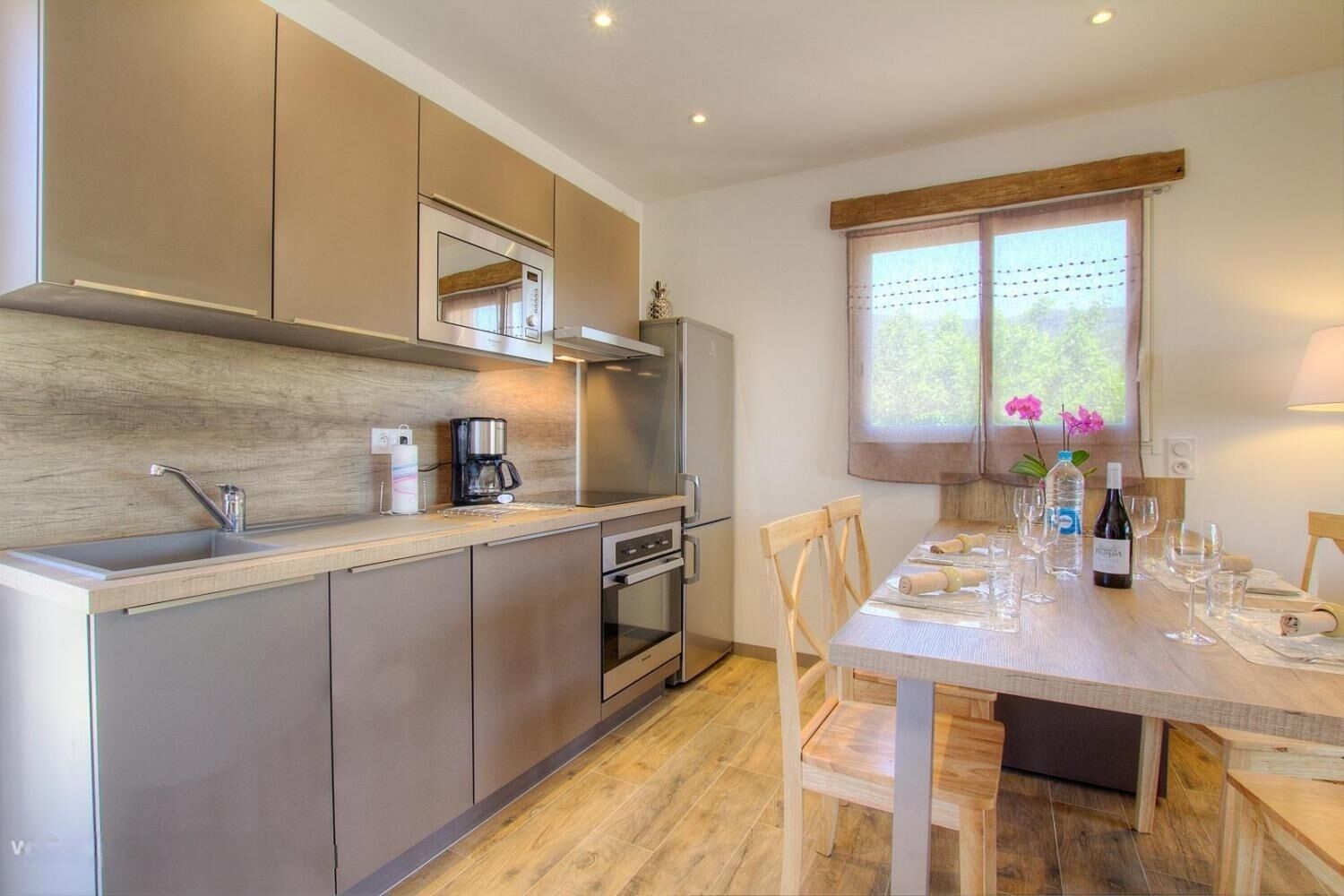 Een moderne keuken met een ingebouwde oven, magnetron en koffiezetapparaat. Er staat een eettafel met stoelen en een raam met gordijnen.