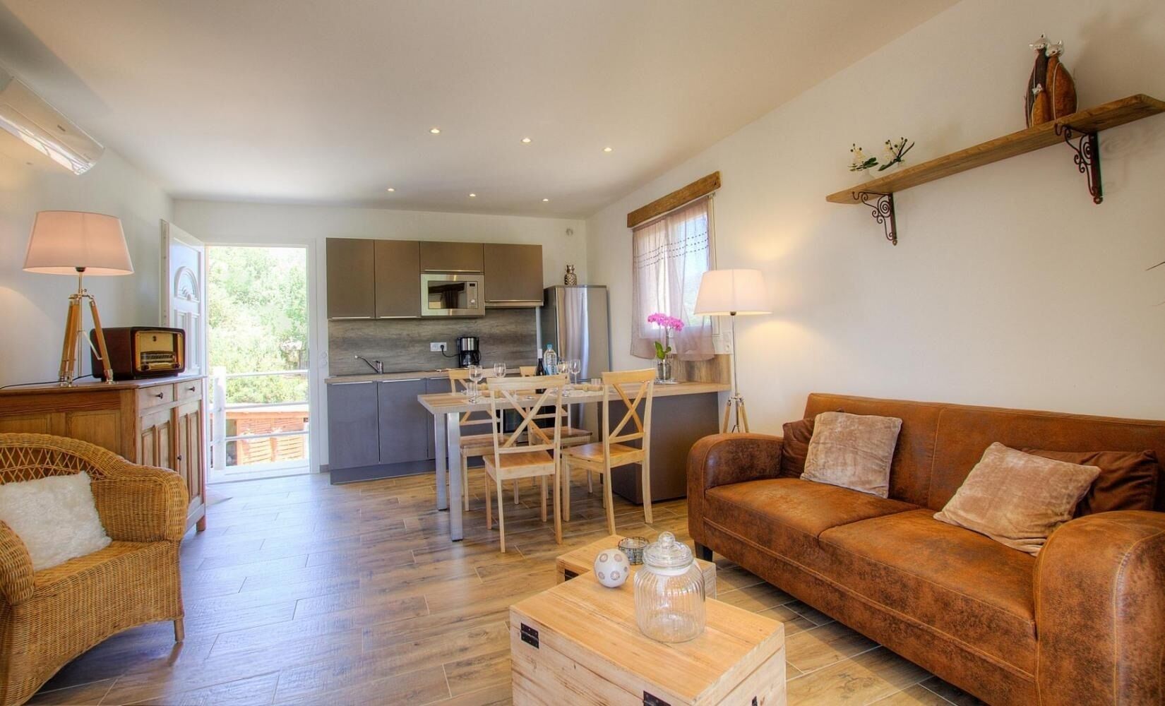 A spacious living area with a wooden floor, a brown leather sofa, a dining table with chairs, and a kitchen in the background.