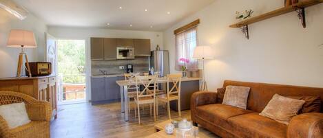 A spacious living area with a wooden floor, a brown leather sofa, a dining table with chairs, and a kitchen in the background.