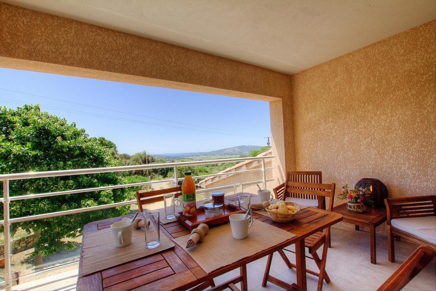 A balcony with a wooden table, chairs, and a view of greenery and hills.