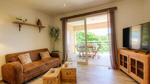 Ein Wohnzimmer mit braunem Ledersofa, einem Holztisch und einem Fernseher auf einer Holzkommode. Ein Balkon mit Tisch und Stühlen ist ebenfalls vorhanden.