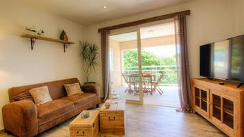 A living room with a brown leather sofa, wooden coffee table, and a TV on a wooden cabinet. There is a balcony with a table and chairs.