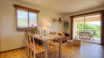 A dining area with a wooden table and chairs, a sofa, a lamp, and a view of greenery through large windows.