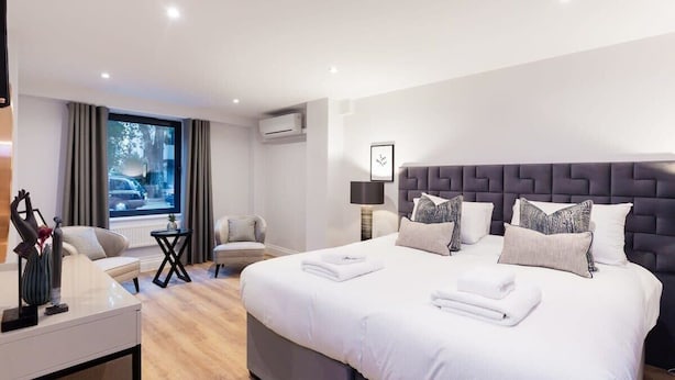 A modern hotel room with a large bed, a desk, a chair, and a view of the street through the window. - Elegant 3BD Apartment in Earls Court (London)