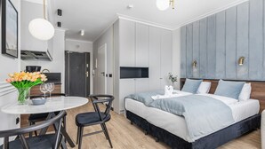 A modern bedroom with a bed, bedside tables, a desk with chairs, a dining area, and a kitchen area.