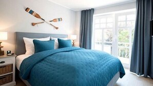 A bedroom with a large bed, a nightstand, a lamp, a wall-mounted paddleboard, and a window with curtains. - Silbermöwe im Lindenhof - designer apartment in country house style (Wangerooge)