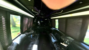 A train interior with black leather seating, a central aisle, and windows providing views of greenery outside. - Limo Party Tailgate Bus (State College)