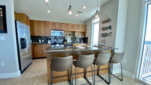 Una cocina moderna con una isla central, taburetes de bar, electrodomésticos de acero inoxidable y un amplio ventanal que proporciona luz natural.