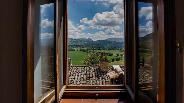 Vista da una finestra con tetto in tegole su un paesaggio verdeggiante, con colline e cielo sereno.